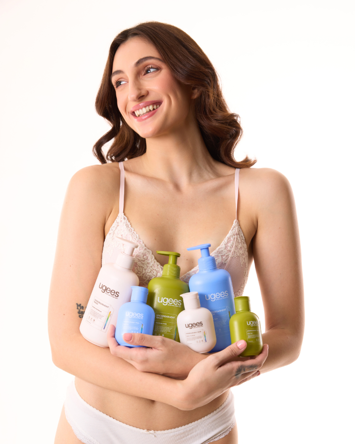 A smiling woman in a white lace camisole and white underwear holds a group of six Uges skincare bottles in her hands against a solid white background.