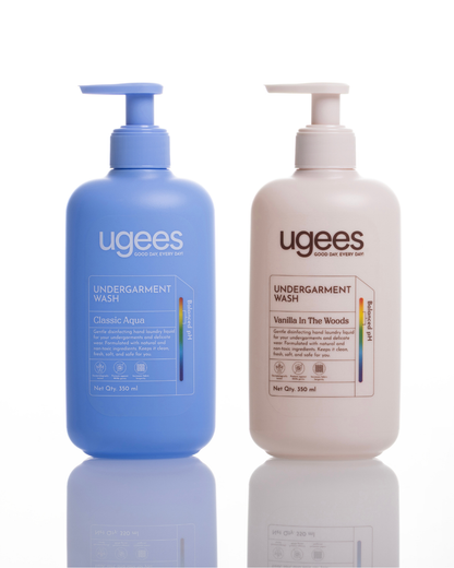 Two Ugees Undergarment Wash bottles on a white reflective background. The left bottle is Classic Aquarius in blue, and the right bottle is Vanilla In The Woods in a warm off-white/cream color.