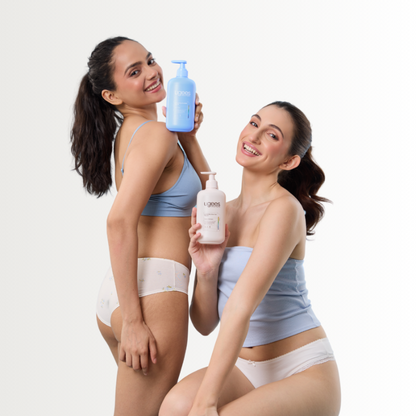 Two women in light blue tank tops and white underwear pose together against a white studio background, each holding a Ugees undergarment wash bottle. One bottle is blue and the other is white with a beige pump.