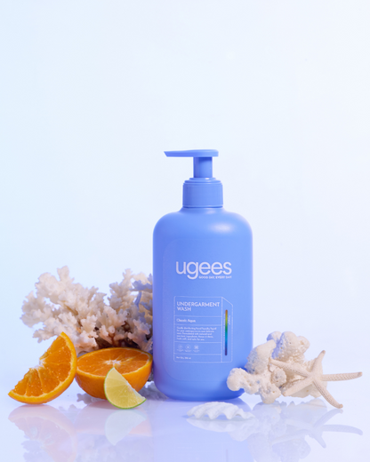 A bottle of ugees undergarment Wash in a light blue pump bottle, styled with citrus slices, starfish, and coral on a reflective white surface against a light blue background.