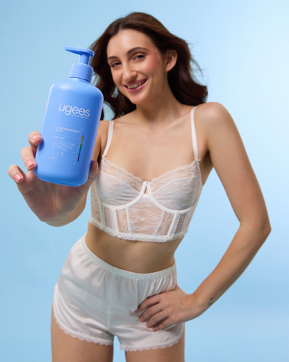 A woman in white lingerie holds up a blue pump bottle of ugees lingerie wash against a light blue studio background.