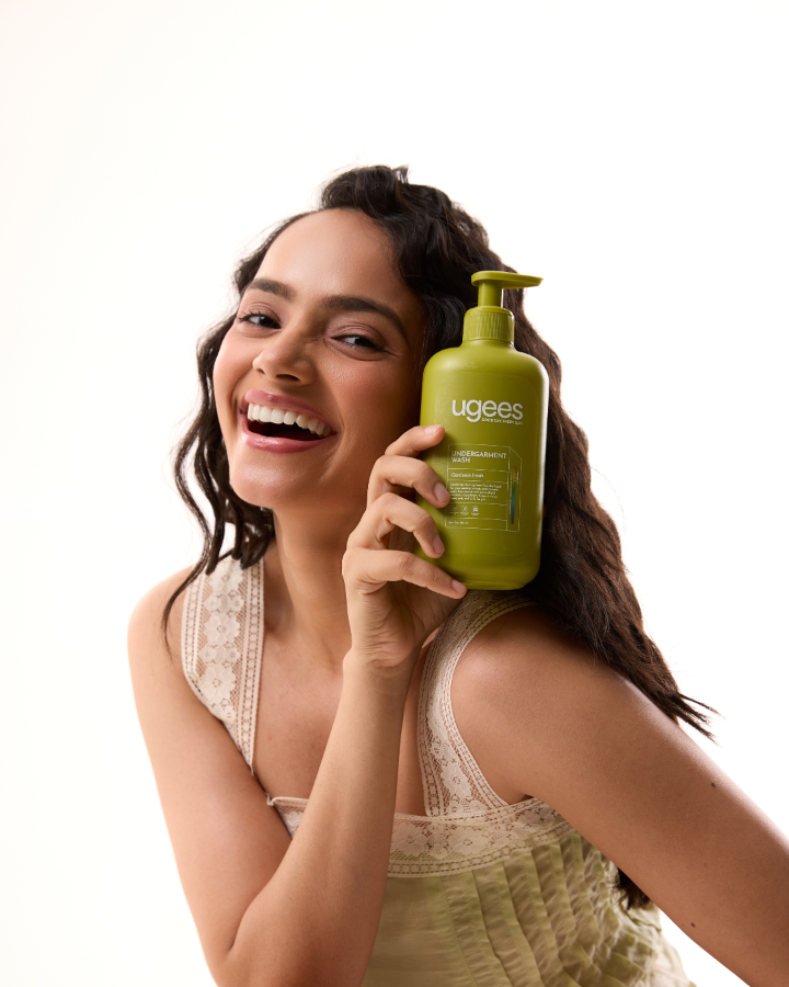 A smiling woman with dark curly hair holds a green pump bottle of ugees undergarment wash  against a clean white background.