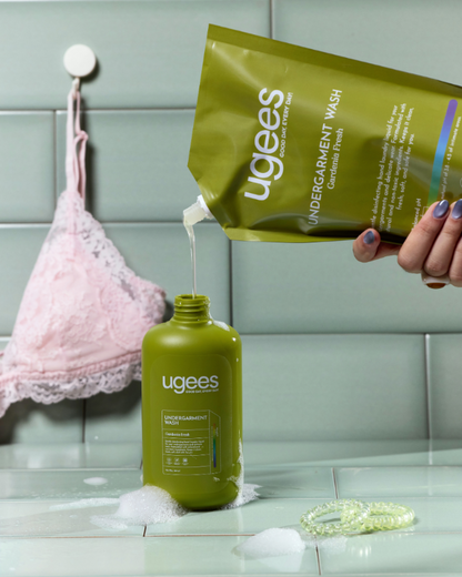 A hand pours clear underarm wash from a green Ugees pouch into a matching green bottle on a bathroom counter, with a pink lace bra visible in the background.