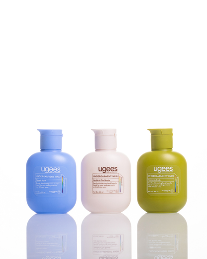 Three Ugees Underwear, swimwear and delicates care bottles lined up on a reflective white surface against a white background. The bottles are light blue, off-white, and olive green, with white caps.