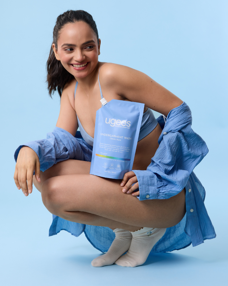 A woman crouches against a light blue studio background, holding a blue refill pack of Ugees Instant Laundry Pack. She is wearing a light blue camisole, an unbuttoned blue shirt draped off her shoulders, and white socks.