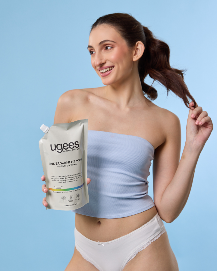 A woman with her hair in a ponytail holds a pouch of ugees Undergarment Wash against a solid light blue background. She is wearing a light blue strapless top and white underwear.