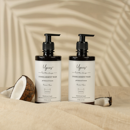 Two pump bottles of Ugees undergarment liquid wash, with a coconut and coconut shells in the background, presented on a neutral backdrop.