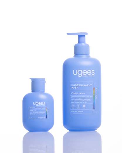 Two blue Ugees Undergarment Wash bottles, one larger pump bottle and one smaller cap bottle, on a white background with a soft reflection below.