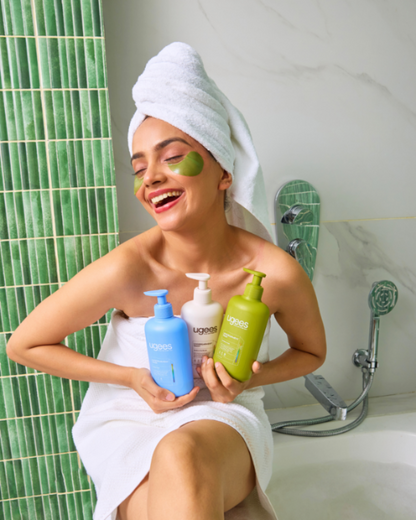 A woman wrapped in a white towel with a white towel turban sits in a bathtub, holding three ugees undergarment wash bottles from the brand. She has a green eye mask .