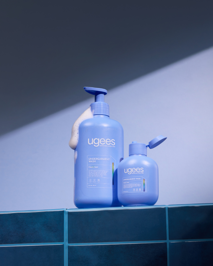 Two Ugees shower products in a blue color scheme, displayed on a dark teal tiled ledge against a blue-gray background with dramatic diagonal lighting.
