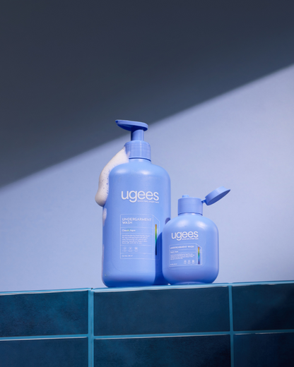 Two Ugees shower products in a blue color scheme, displayed on a dark teal tiled ledge against a blue-gray background with dramatic diagonal lighting.