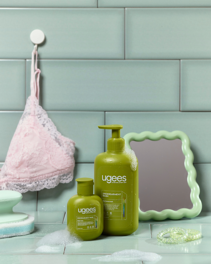 Two green Ugees undergarment wash bottles on a tiled bathroom counter with a pink bra hanging on a hook, a small mirror, and soap residue.