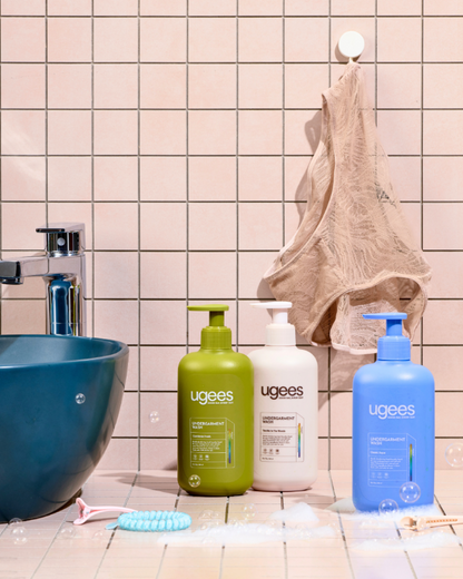 Three Ugees body wash bottles in green, white, and blue, arranged on a bathroom counter next to a blue sink, with a beige underwear hanging on a hook in the background.