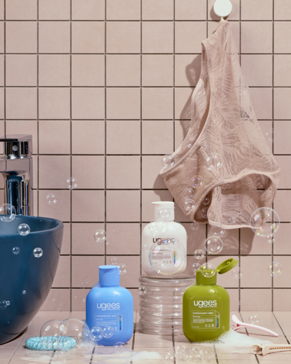 Three Ugees underwear wash bottles in blue, white, and green are arranged on a tiled bathroom counter next to a sink, with a beige underwear hanging on a hook behind them and floating bubbles around the scene.