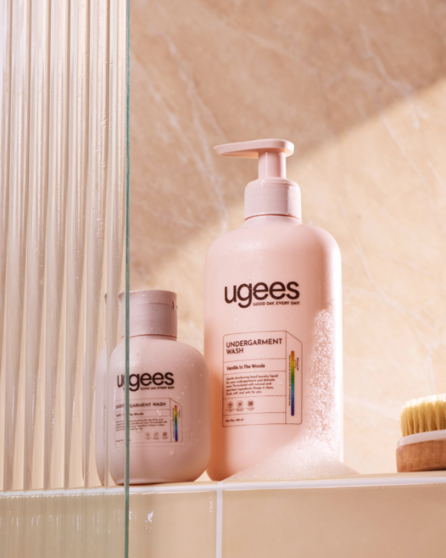 Two Ugees Undergarment Wash bottles in a warm beige bathroom setting, with one larger pump bottle and one smaller bottle on a shelf next to a glass divider.